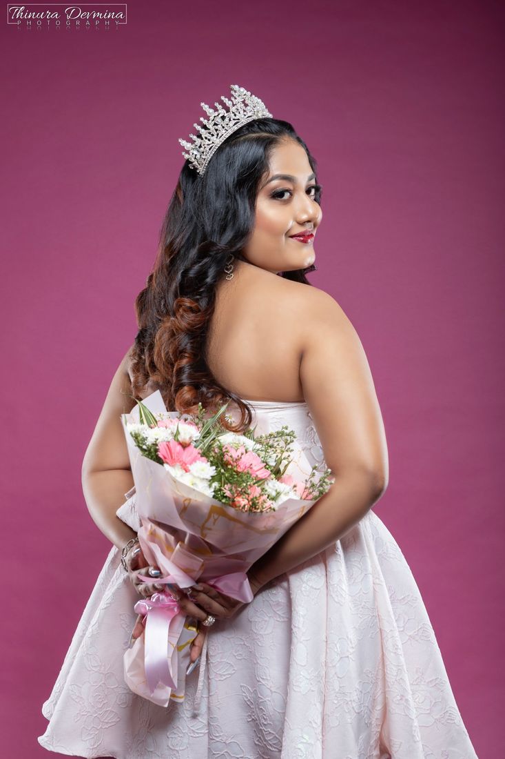 A crowned birthday girl glancing over her shoulder holding a bouquet behind her back